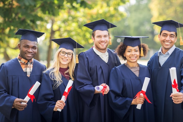 education, graduation and people concept - group of happy international students in mortar boards and bachelor gowns with diplomas