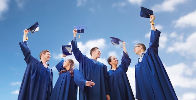 education, graduation and people concept - group of smiling students in gowns waving mortarboards over blue sky and clouds background