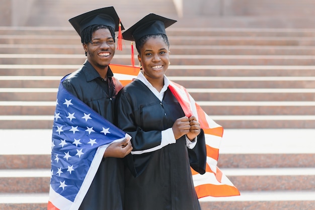 Концепция образования, выпуска и людей - счастливые иностранные студенты в минометных досках и бакалаврских халатах с американским флагом