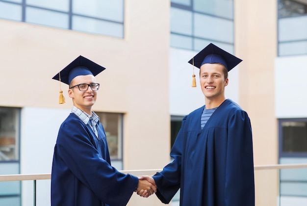 education, graduation and people concept - smiling students in mortarboards and gowns shaking hands outdoors