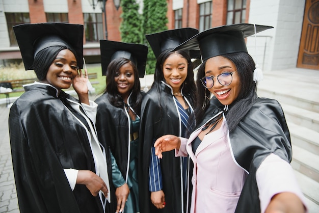 Education, graduation, technology and people concept - group of\
happy international students in mortar boards and bachelor gowns\
with diplomas taking selfie by smartphone outdoors