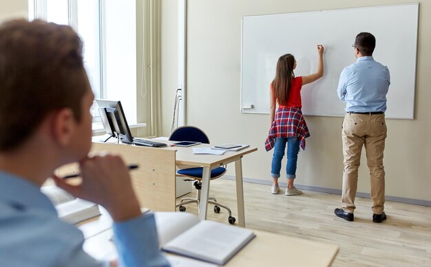 Foto istruzione, scuola superiore, apprendimento e concetto di persone - studentessa che scrive qualcosa su una lavagna bianca vuota e insegnante in classe