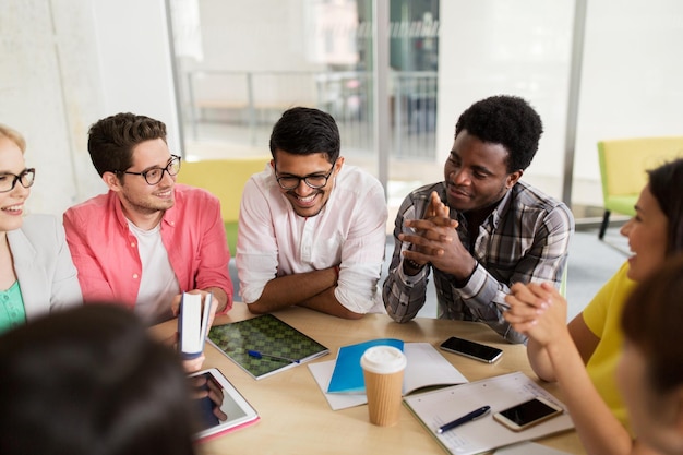 Photo education, high school, people and technology concept - group of international students sitting at table with tablet pc computer, smartphones and notebooks and talking at university