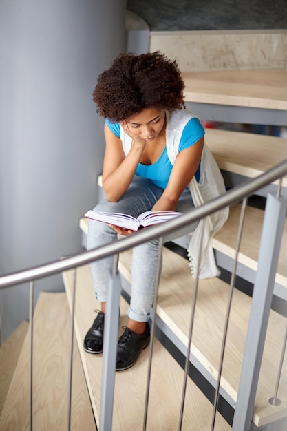 Foto educazione, scuola superiore, università, apprendimento e concetto di persone - ragazza studentessa afroamericana che legge libro seduto sulle scale in biblioteca