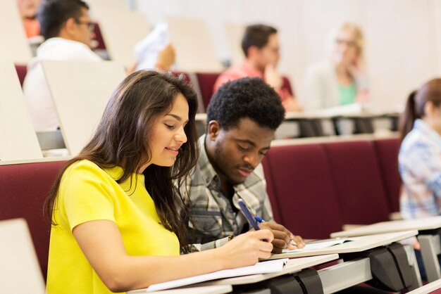 education, high school, university, learning and people concept - group of international students with notebooks writing in lecture hall