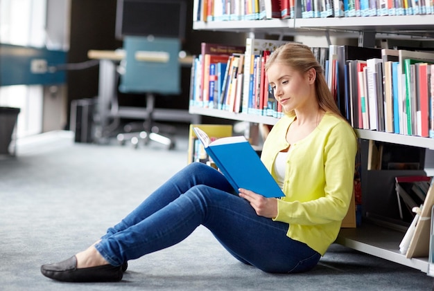 образование, средняя школа, университет, обучение и концепция людей - улыбающаяся студентка читает книгу, сидя на полу в библиотеке