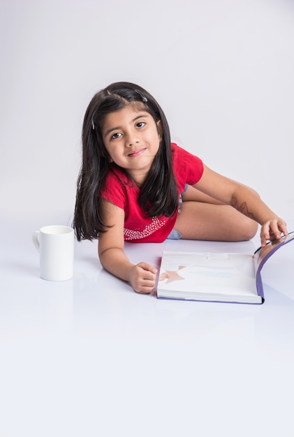 Education at home concept - Cute little Indian or Asian girl studying with pile of books and coffee mug while sitting on the floor at home. Isolated over white background