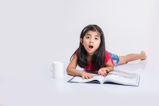 Education at home concept - Cute little Indian or Asian girl studying with pile of books and coffee mug while sitting on the floor at home. Isolated over white background