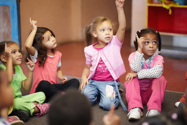 Фото Детский сад образования и дети, задающие вопрос с поднятыми руками, сидя на полу в классе для развития ребенка школьное обучение или любопытство с детьми в классе, чтобы учиться для роста