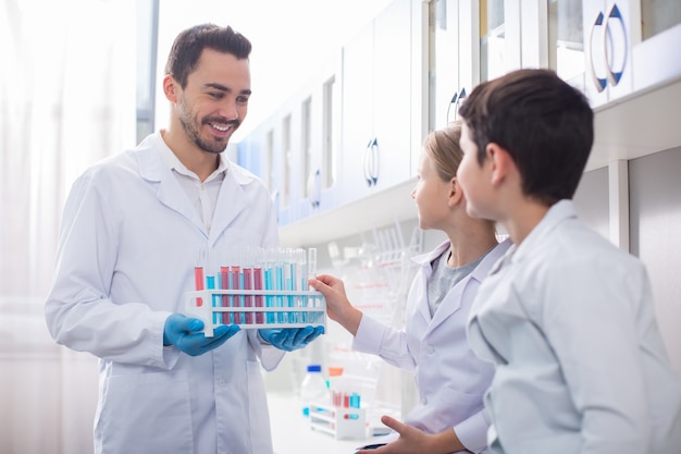 Education at lab. Bearded cheerful man holding stand while girl taking vial from there and kids staring at him