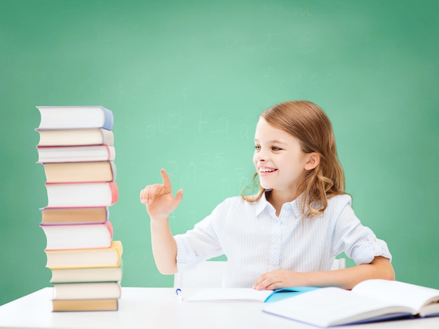 education, people, children and school concept - happy student girl sitting at table and counting books over green chalk board background