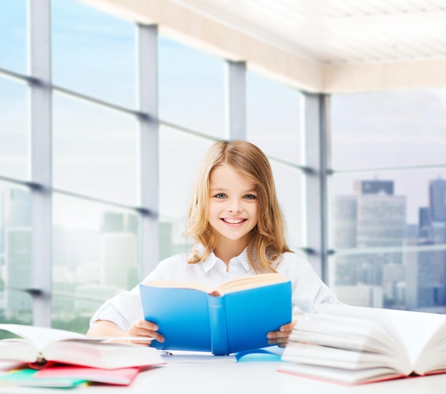 education, people, children and school concept - little student girl sitting at table with books and writing in notebook over classroom background