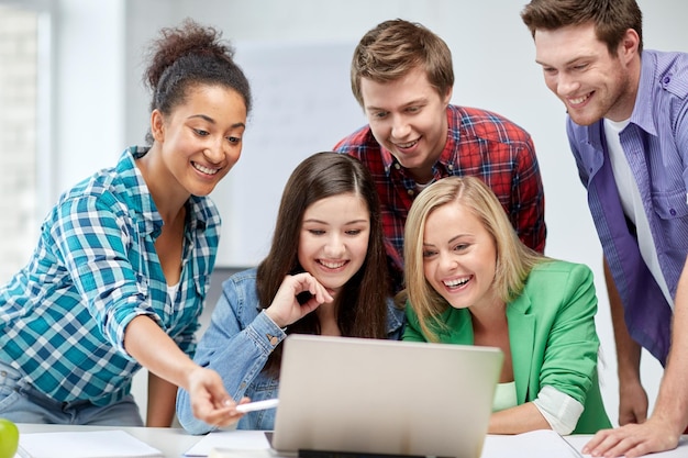 Education, people, friendship, technology and learning concept\
- group of happy international high school students or classmates\
with laptop computer in classroom