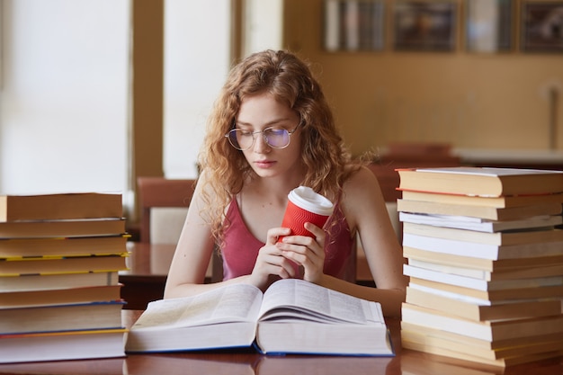 Education and school concept. Clever female student girl with coffee in hand, wearing glasses and casual outfit, looks concentrated, sitting at desk in college library with stack books reading.