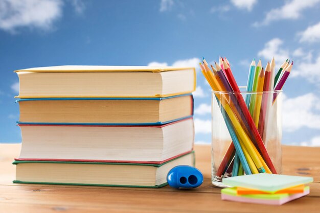 교육, 학교, 창의성 및 개체 개념 - 푸른 하늘과 구름 배경 위에 있는 나무 테이블에 책, 스티커 및 샤프너가 있는 크레용 또는 색연필을 닫습니다.