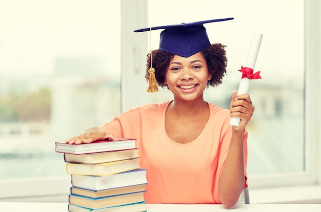 education, school, knowledge, graduation and people concept - happy smiling african american student girl in bachelor cap with books and diploma sitting at table at home