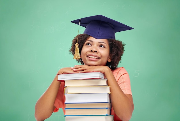 Photo education, school, knowledge and people concept - happy smiling african american student girl in bachelor cap with books sitting at table and dreaming over green chalk board background