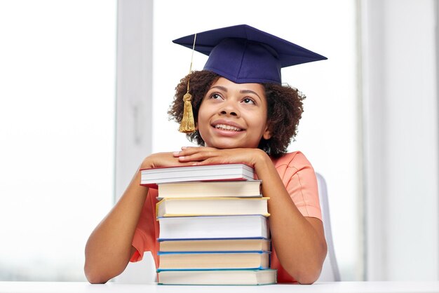 education, school, knowledge and people concept - happy smiling african american student girl in bachelor cap with books sitting at table and dreaming at home