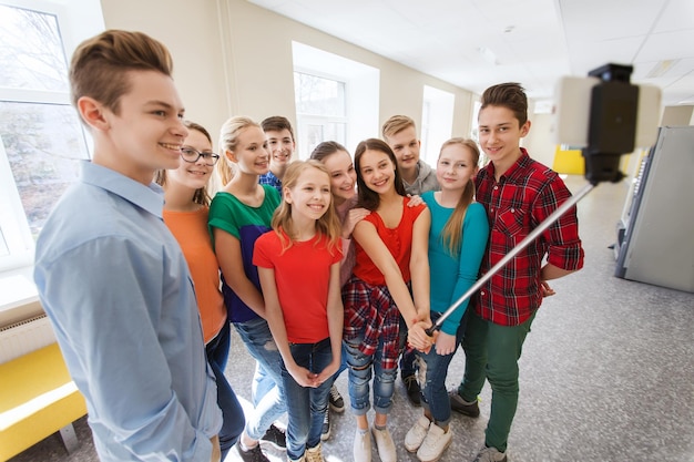 教育、学校、技術、人々 のコンセプト - 廊下でスマート フォンの自撮り棒で写真を撮る幸せな笑顔の学生のグループ