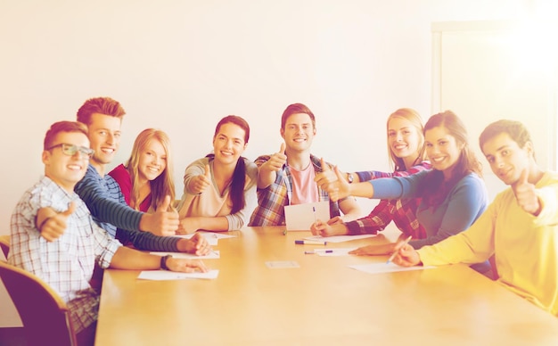 образование, работа в команде, жест и концепция людей - улыбающиеся студенты с бумагами, показывающие большие пальцы вверх и сидящие за столом