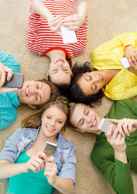 education, technology and happiness concept - group of young smiling people lying down on floor in circle with smartphones