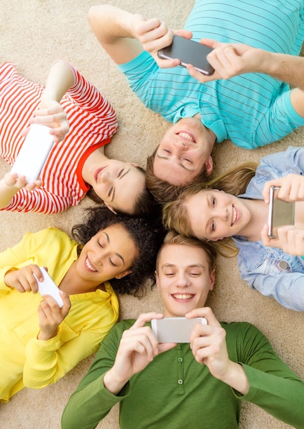 education, technology and happiness concept - group of young smiling people lying down on floor in circle with smartphones