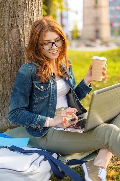 教育、技術、インターネットの概念-ラップトップコンピューターを備えた眼鏡で赤毛のティーンエイジャーを笑顔にし、コーヒーやお茶を奪う