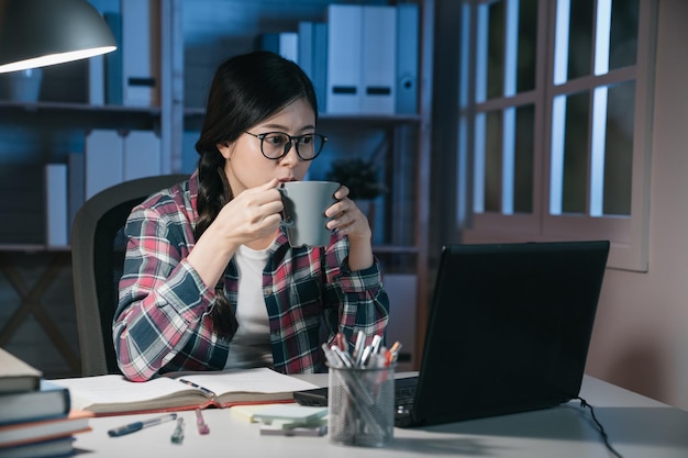 教育技術と人々 の概念。ラップトップ コンピューターの画面を見つめ、夜の家でコーヒーを飲む眼鏡をかけているアジアの日本人学生の女の子。勤勉な女性はお茶を楽しみます。