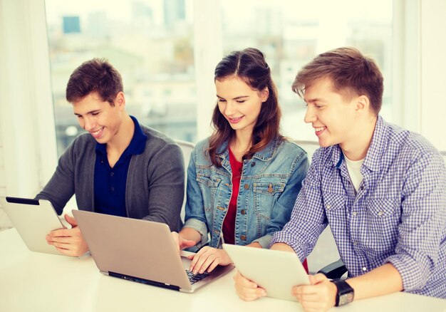 education, technology, school and internet concept - three smiling students with laptop and tablet pc at school