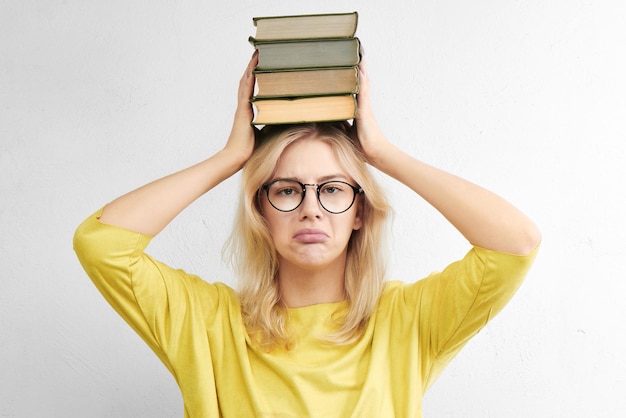 Educational concept. European student girl in glasses holds heavy textbooks on head with sad face and cry, under stress on a white background