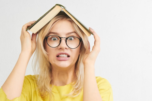 Educational concept. European student girl in round glasses and yellow clothes holds a textbook on head, stress from studying. Portrait on a white studio background