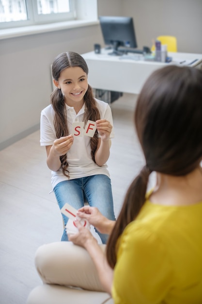 Educational game. Smiling smart girl with letter cards in hands and woman psychologist sitting with back to camera