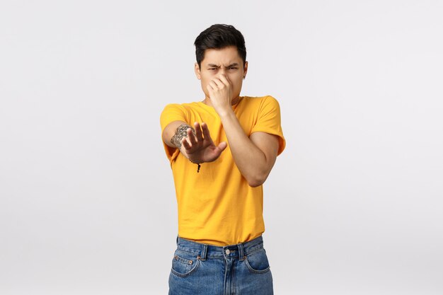 Eeew stinky. Disgusted asian male in yellow t-shirt, cover nose from aversion, smell something awful, blame or accuse person reek, standing white wall displeased