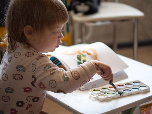 Een 2-jarig meisje zit aan een tafel en leert tekenen met veelkleurige aquarellen Kindercreativiteit Kinderhobby's