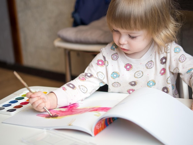 Een 2-jarig meisje zit aan een tafel en leert tekenen met veelkleurige aquarellen Kindercreativiteit Kinderhobby's