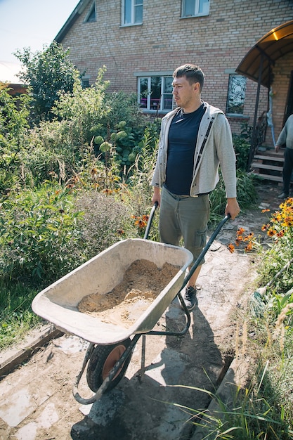 Een 30-jarige blanke man draagt een tuinkruiwagen voor het laden van zand om de paden met beton te vullen in zijn zomerhuisje