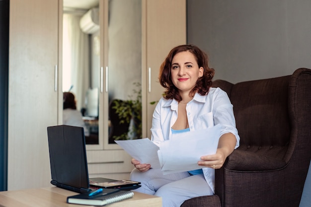 Een 30-jarige vrouw met donker haar en een wit overhemd zit in een fauteuil aan een tafeltje met een laptop, houdt papier in haar handen en werkt op afstand in haar kantoor aan huis.
