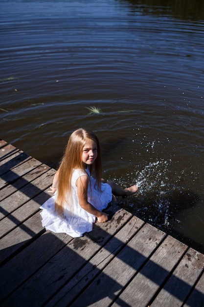 Een 7-jarig meisje met lang blond haar bij het meer zit op een clutch met benen in het water. ze spettert met haar voeten in het meer. barefoot meisje in een witte jurk met lang haar.