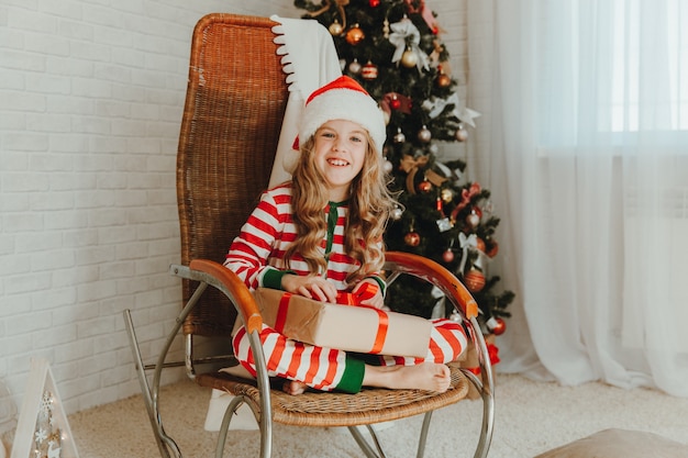 Een 9-jarig meisje met een geschenk van de kerstman en zittend op een schommelstoel met slingers op de achtergrond. Kersttijd. Gelukkig nieuwjaar!