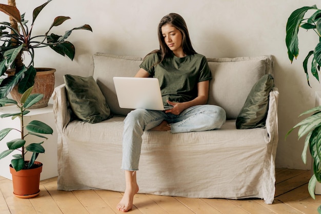 Een aantrekkelijk jong brunette meisje dat op een bank zit gebruikt een laptop om online op afstand te werken