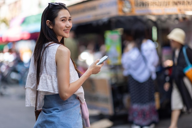 Een aantrekkelijke jonge Aziatische toerist loopt op straat in een winkelmarkt in Thailand. Ze gebruikt een smartphone om een bericht te sturen en belt iemand op de mobiele telefoon.