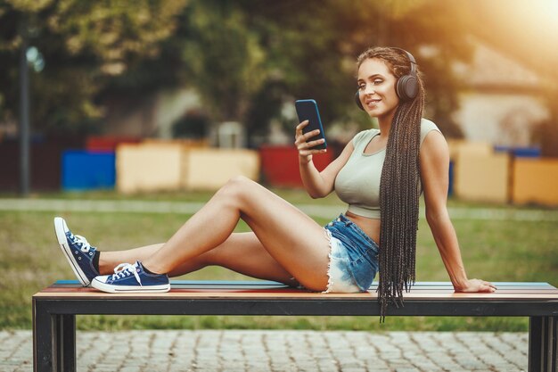 Een aantrekkelijke jonge blanke vrouw met afro gevlochten haar ondergedompeld in gedachten met een koptelefoon op haar hoofd zit op de kleurrijke bank te kijken naar de smartphone en de muziek te luisteren.