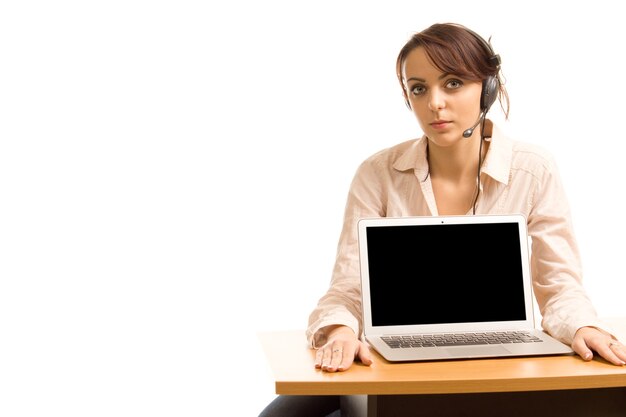 Een aantrekkelijke jonge call centreexploitant zit aan een bureau in een hoofdtelefoon met het lege scherm van haar laptop