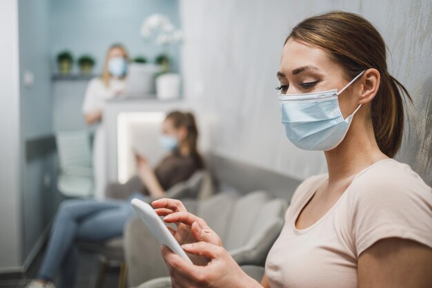 Een aantrekkelijke jonge vrouw die een gezichtsmasker draagt en haar mobiele telefoon gebruikt terwijl ze op een stoel zit in de wachtkamer op het kantoor van de tandarts.