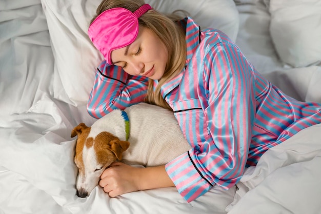 Een aantrekkelijke jonge vrouw in pyjama houdt een hond vast terwijl ze op een bed ligt