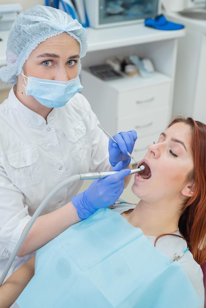 Een aantrekkelijke lachende vrouwelijke arts in wit uniform onderzoekt een roodharige vrouwelijke patiënt Tandarts produceert traktaties tanden met een boor