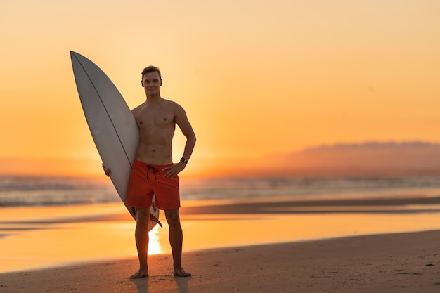 Een aantrekkelijke man die op de kust staat en een surfplank vasthoudt bij oranje zonsondergang