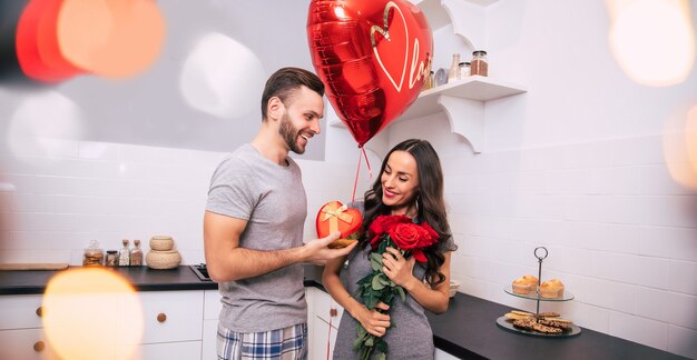 Een aantrekkelijke man en een knappe vrouw in huiskleding staan in de keuken en glimlachen terwijl ze elkaar cadeautjes geven op Valentijnsdag.
