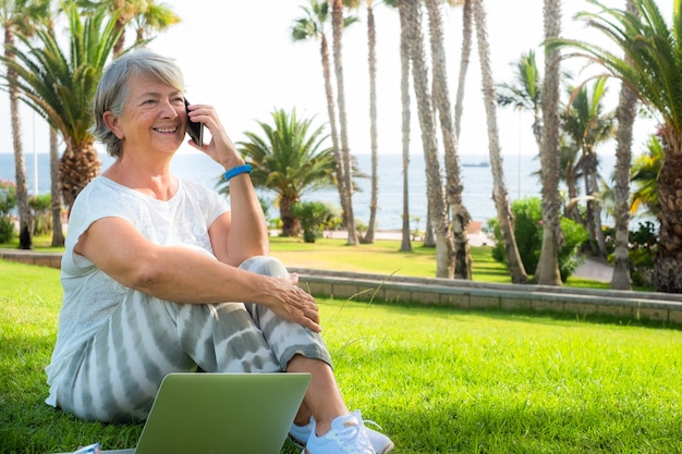 Een aantrekkelijke senior vrouw zittend op het gazon met laptop met behulp van de mobiele telefoon