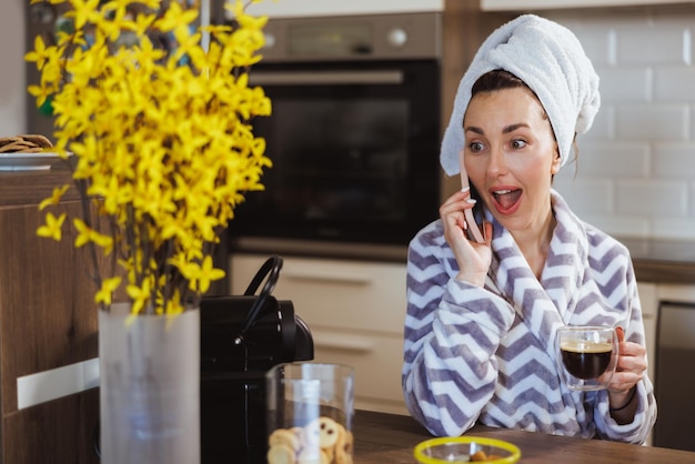 Een aantrekkelijke verraste vrouw in badjas die smartphone gebruikt terwijl ze thuis geniet van koffie in de ochtend.